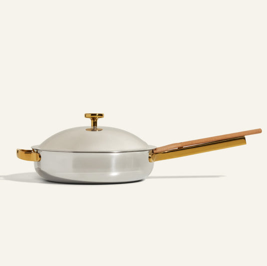 The Titanium Always Pan Pro, shown on a plain light background, is silver with a matching lid, gold-colored handle and knob, wooden accent on the long handle, and features durable no-coating nonstick technology.
