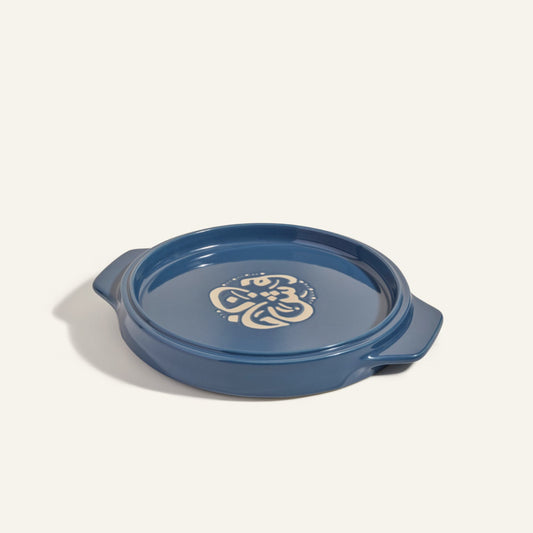 The Flipping Platter is a blue ceramic baking dish with handles and a cream floral design in the center, perfect for serving dishes like tahdig. Shown against a plain light background.