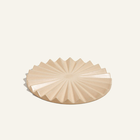 The Pleat Trivet is a beige ceramic plate with a geometric starburst pattern and ridged, angular edges—ideal as a decorative kitchen accessory or unique sunburst trivet, shown on a plain light background.
