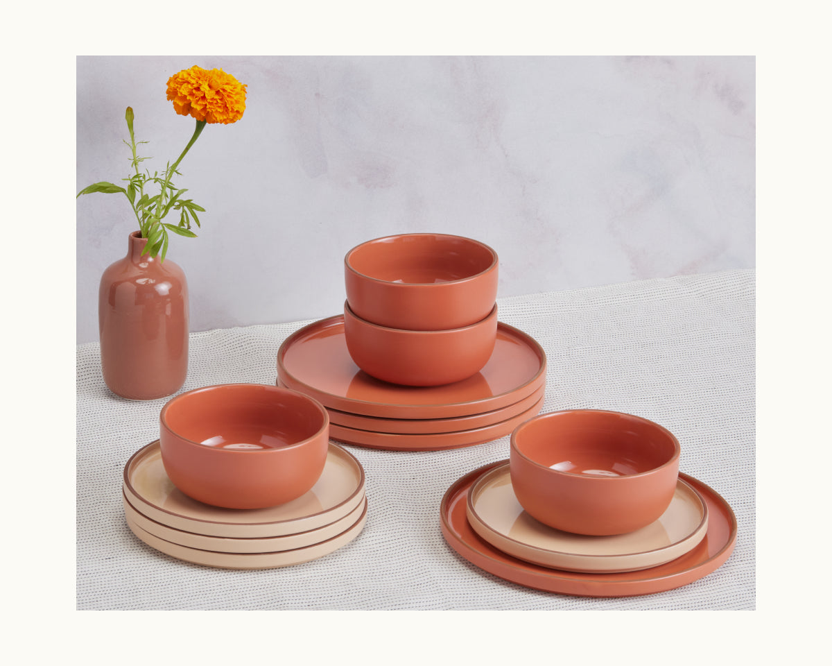 A set of terracotta and beige Soup Bowls, designed to stack, is arranged with matching dishes on a table beside a marigold flower in a coordinating vase.