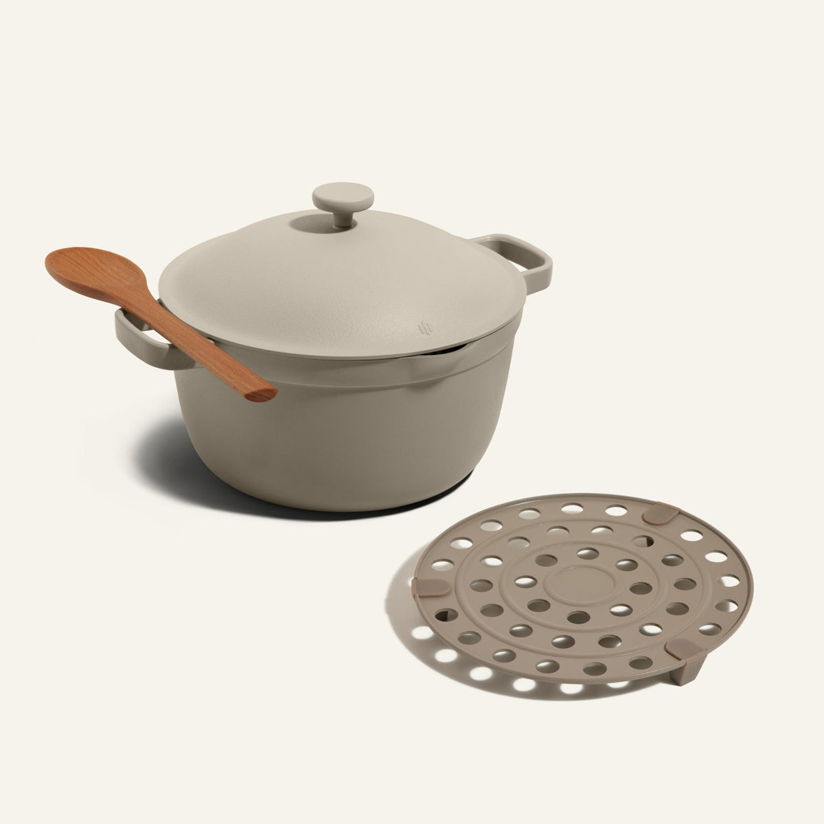 Beige Perfect Pot with lid and wooden spoon on handle, next to the matching beige Perfect Pot Roasting Rack featuring circular holes, set against a plain background.