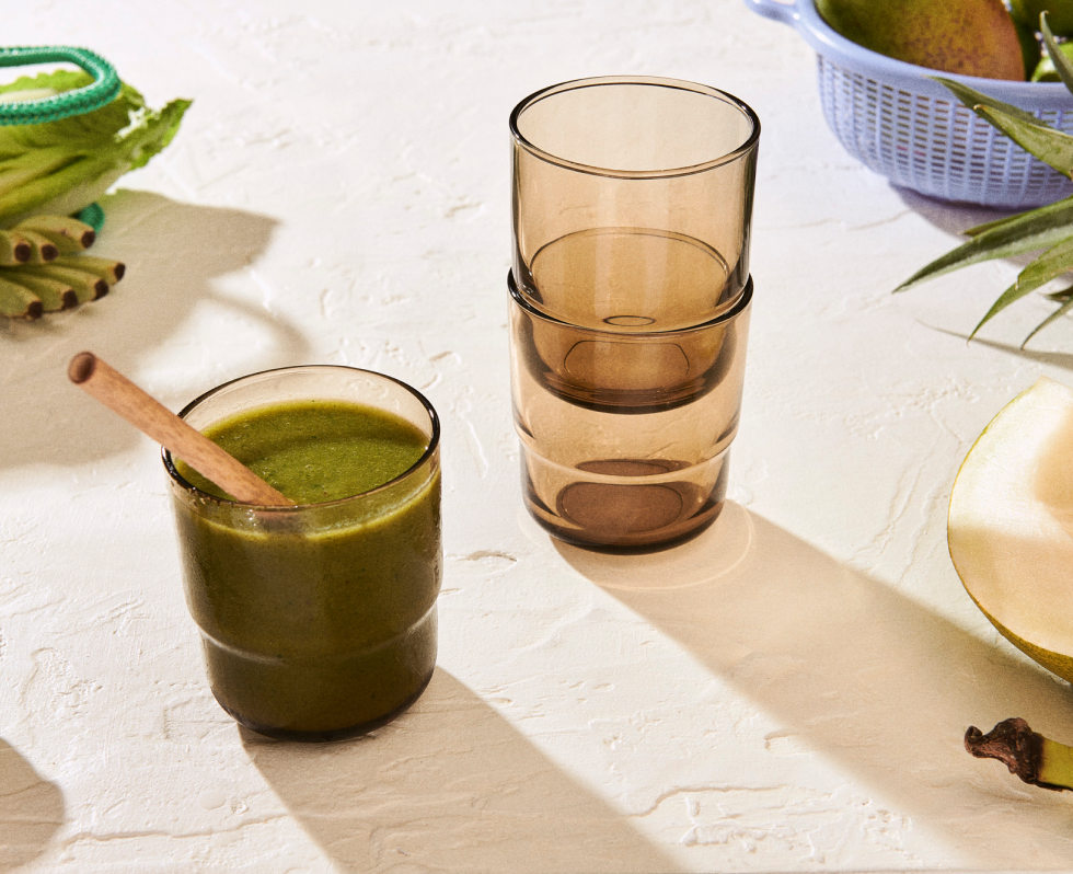 A glass of green smoothie with a wooden spoon sits beside two stacked Short Night + Day Glasses on a textured white surface, with fruit and a blue basket in the background.