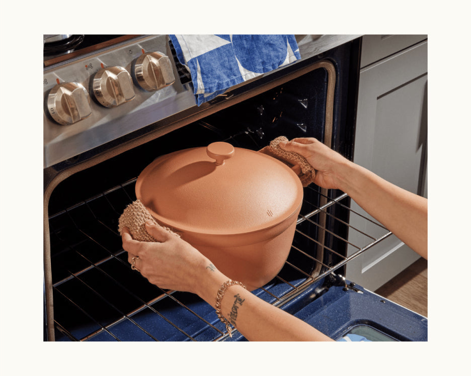 A person uses pot holders to place a lidded Cookware Set pot into an oven, with oven dials and a towel visible in the background.