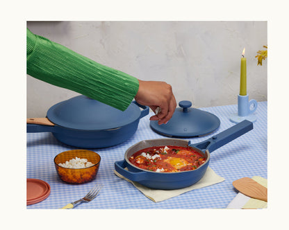 A hand lifts the lid off a blue Cookware Set pot with eggs in tomato sauce, resting on a blue checkered tablecloth beside a bowl of cheese and a lit candle.
