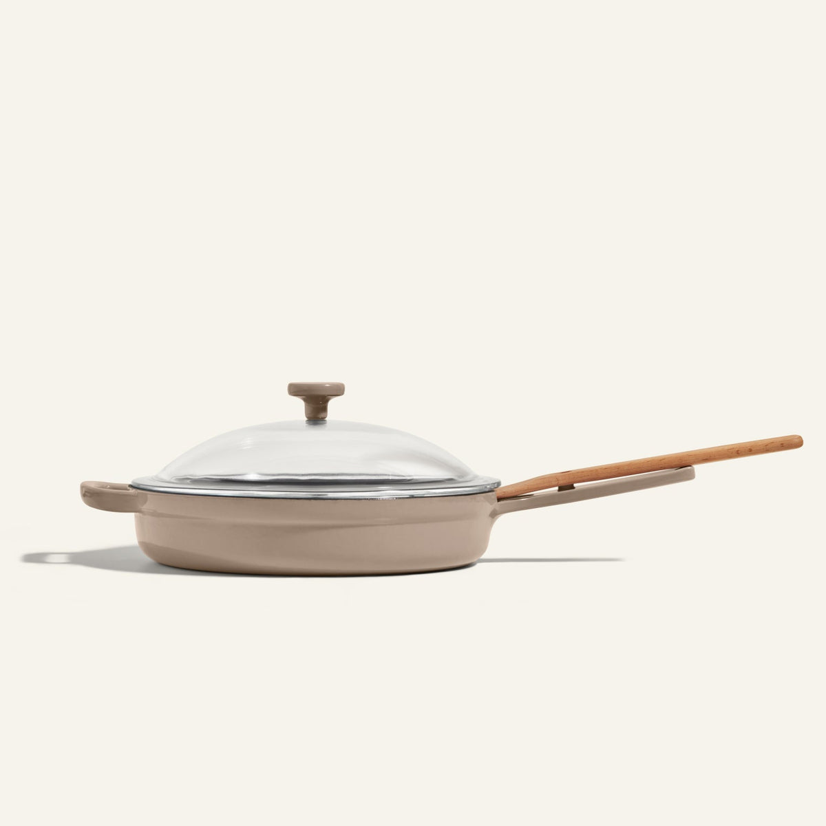 The Cast Iron Always Pan, a beige enameled nonstick frying pan with a wooden handle and glass lid, is photographed against a plain light background.