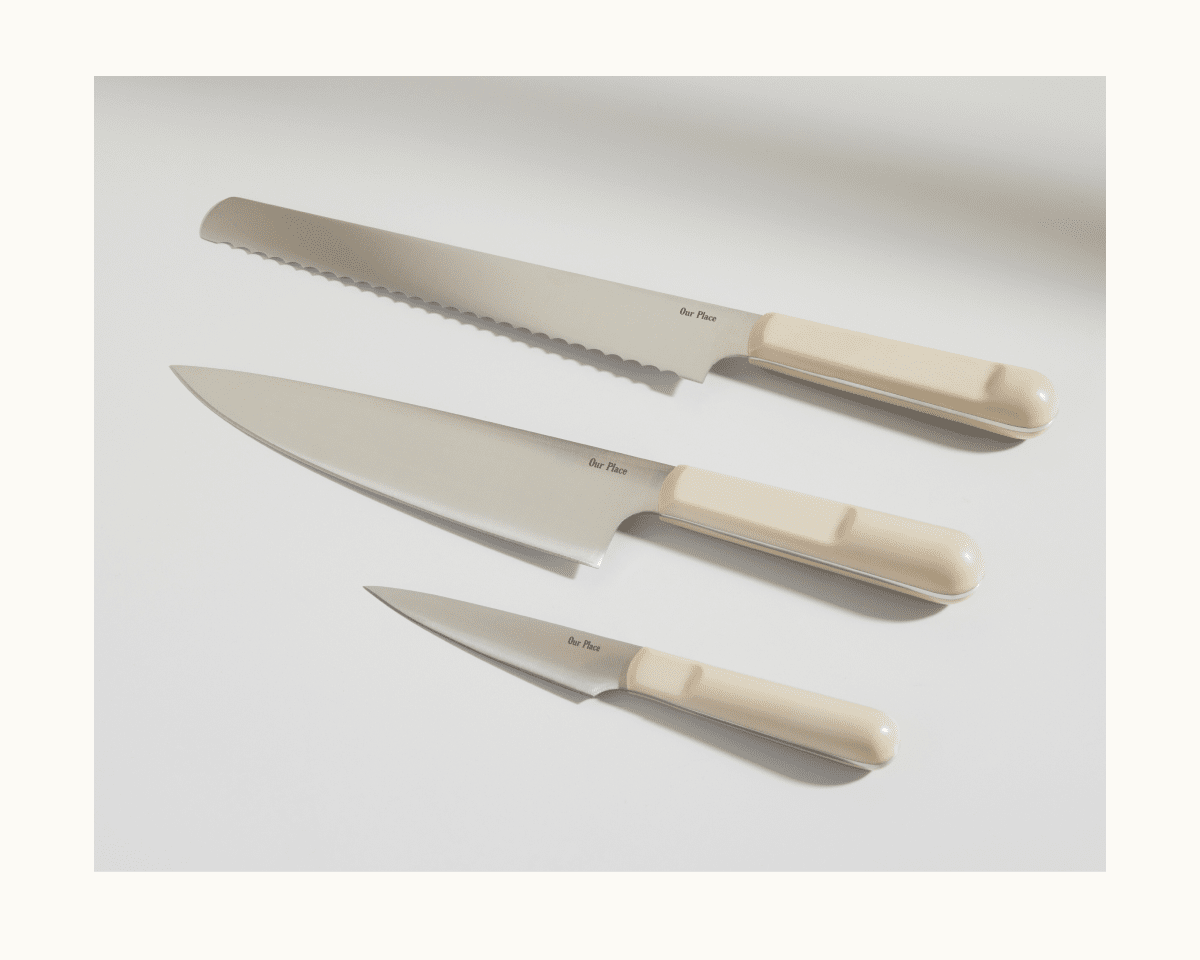 The Everyday Chef’s Knife, featuring a beige handle and crafted from durable German steel, is displayed on a white surface alongside a bread knife and paring knife.