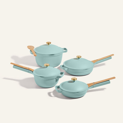 The Cookware Set includes four light blue nonstick pieces inspired by the Always Pan and Perfect Pot: two frying pans, a saucepan, a lidded pot with gold knobs, wooden handles, and a wooden spoon.