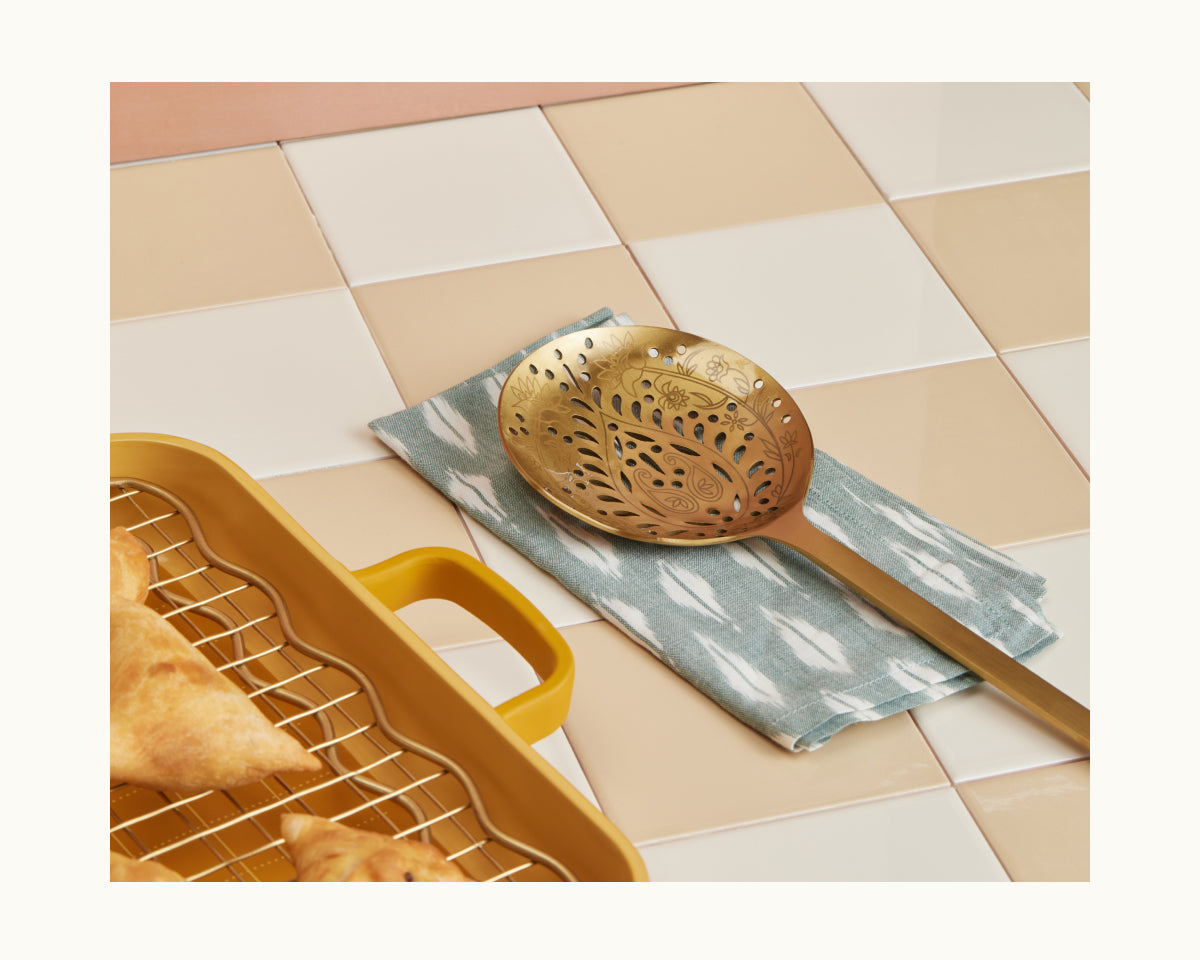 The Golden Fry Set, featuring a decorative brass skimmer spoon, sits on a patterned cloth beside a yellow baking dish filled with pastries—an ideal setup for preparing treats during Diwali celebrations.