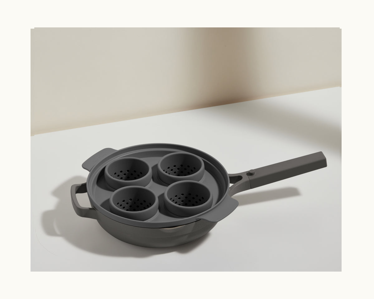 The Egg Poacher, featuring four gray silicone cups and a long handle for easy use, is ideal for making poached eggs. It’s shown on a white surface with a plain background.