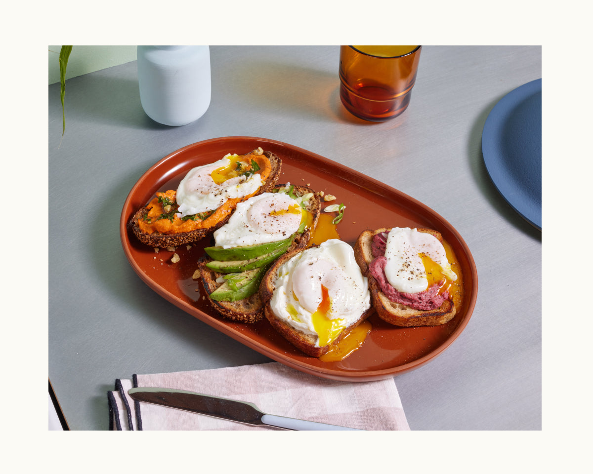 A plate holds four slices of toast, each topped with poached eggs made in the Egg Poacher and various toppings, set beside a knife, napkin, glass, and bottle on the table.