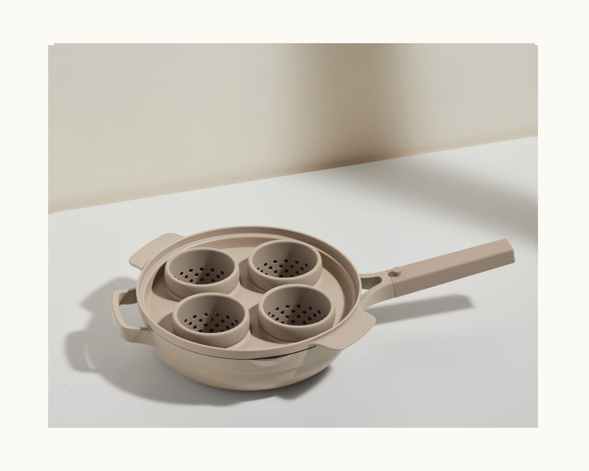 The Egg Poacher, a beige saucepan with four removable perforated inserts for poaching eggs or using silicone cups, rests on a white surface.