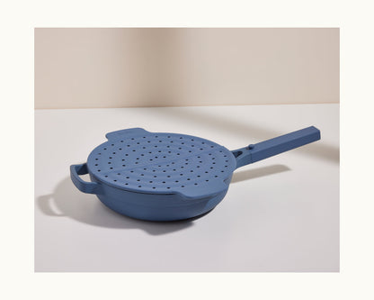 A Fearless Fry pan in blue, featuring a matching perforated lid and long handle for splatter prevention, rests on a white surface against a neutral background.