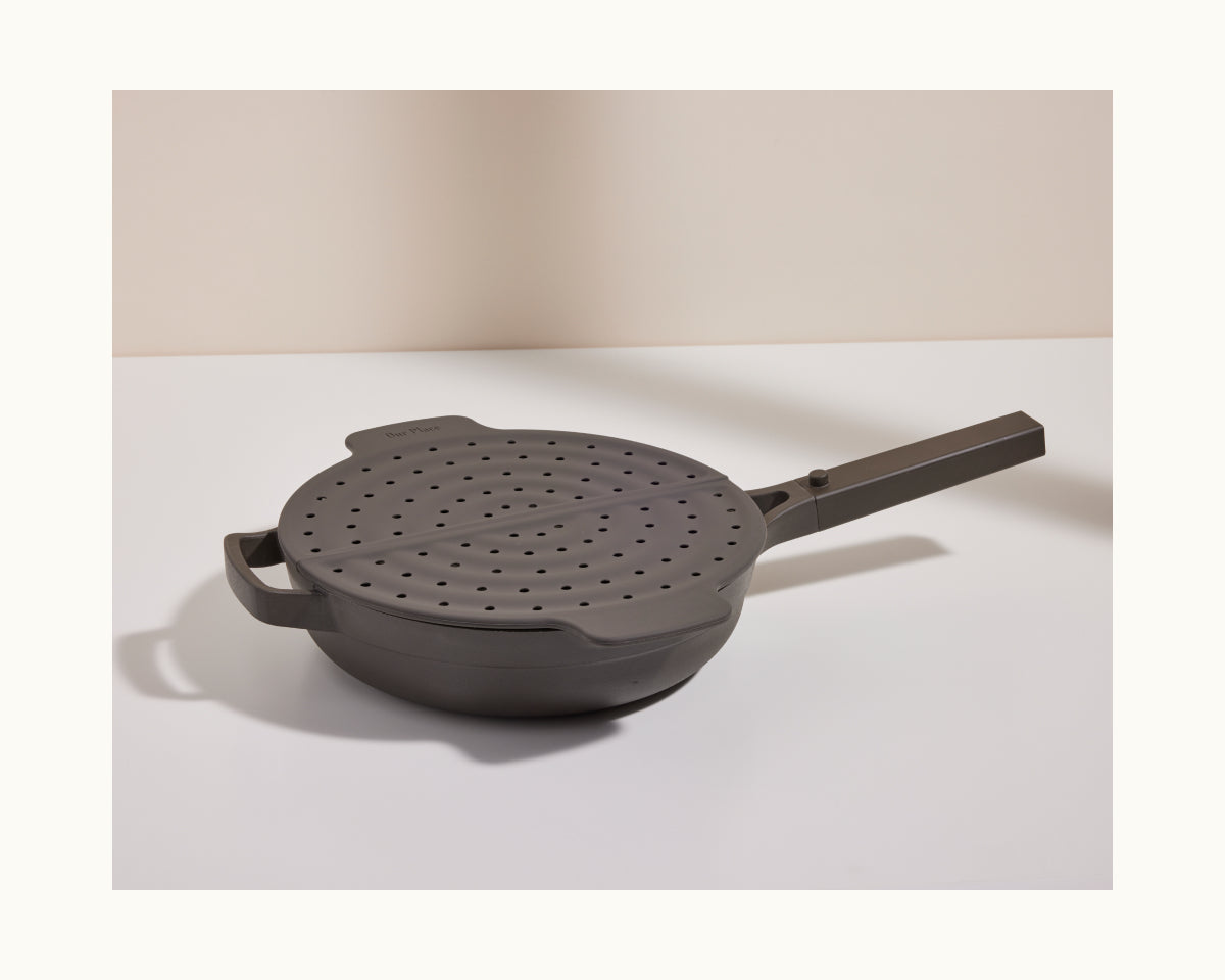 The Fearless Fry, a gray pan with a matching perforated lid and long handle, sits on a white surface, offering stylish splatter prevention against a neutral background.