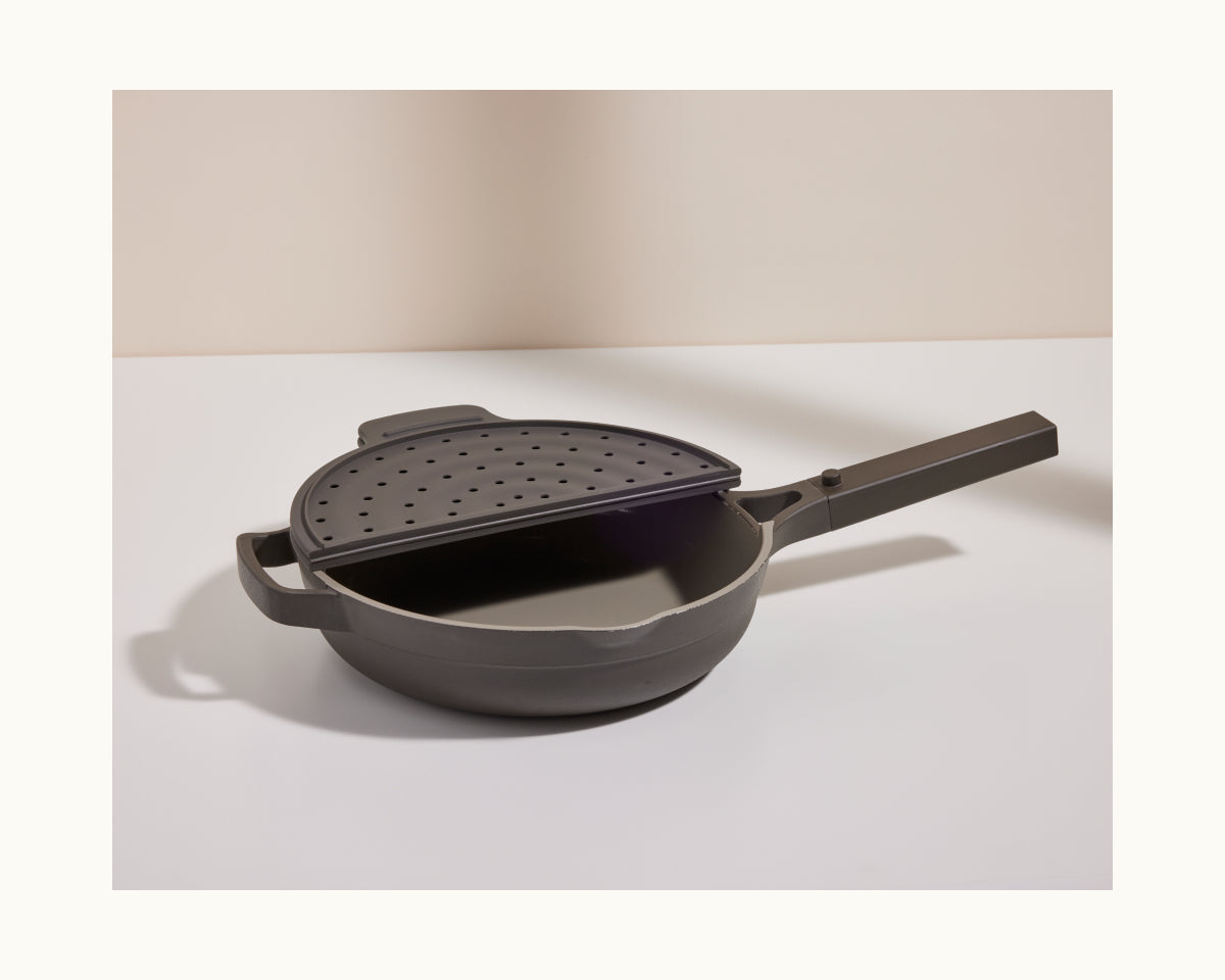 The Fearless Fry pan, in gray nonstick finish, features a long handle, two short handles, and comes with a perforated splatter-prevention lid, shown partially covering the pan on a white surface.