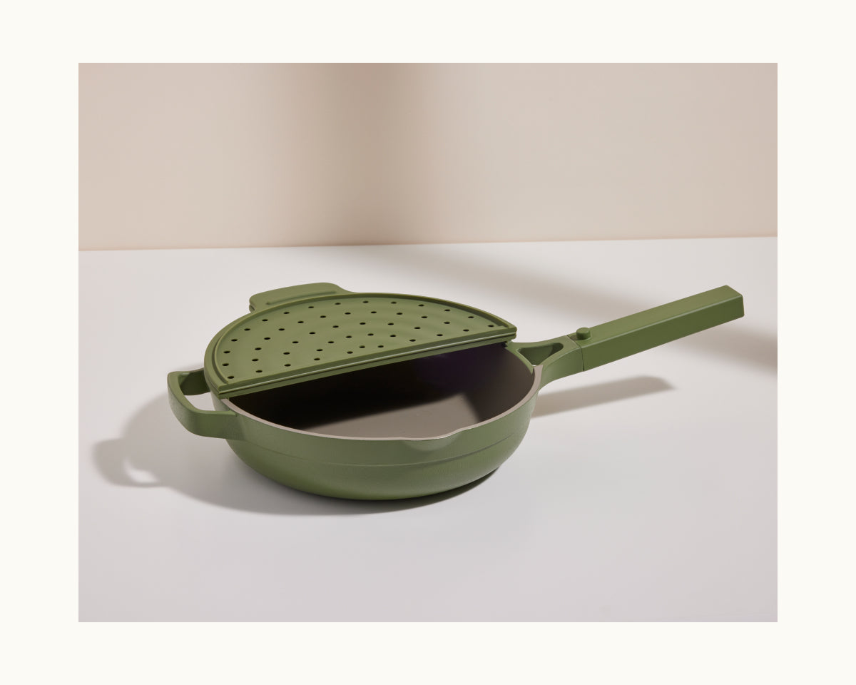 The Fearless Fry, a green nonstick frying pan with a matching handle and perforated splatter-prevention lid inspired by the Always Pan, rests on a white surface against a neutral background.