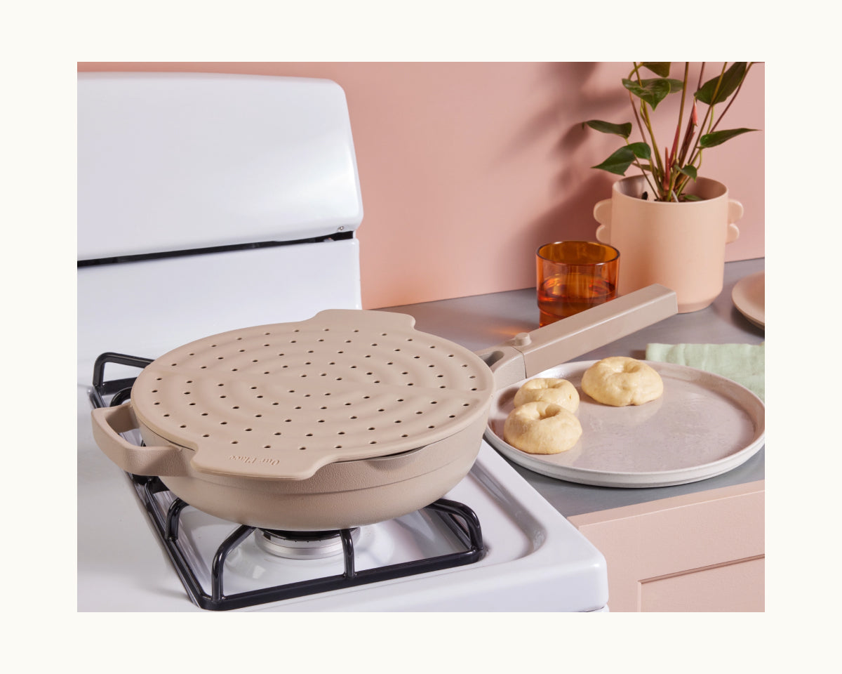 A beige Fearless Fry with a steamer insert sits on a stove beside a plate holding three dough balls, providing splatter protection. A plant and dishes are visible in the background.