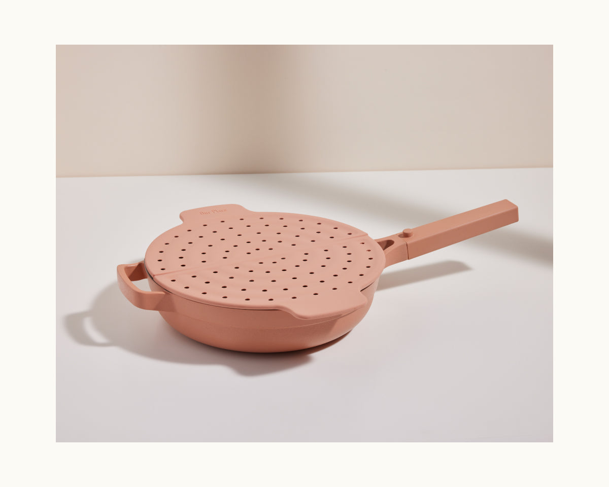 The Fearless Fry, a pink nonstick frying pan inspired by the Always Pan, comes with a matching handled lid featuring multiple steam holes for splatter prevention and sits on a white surface against a neutral background.