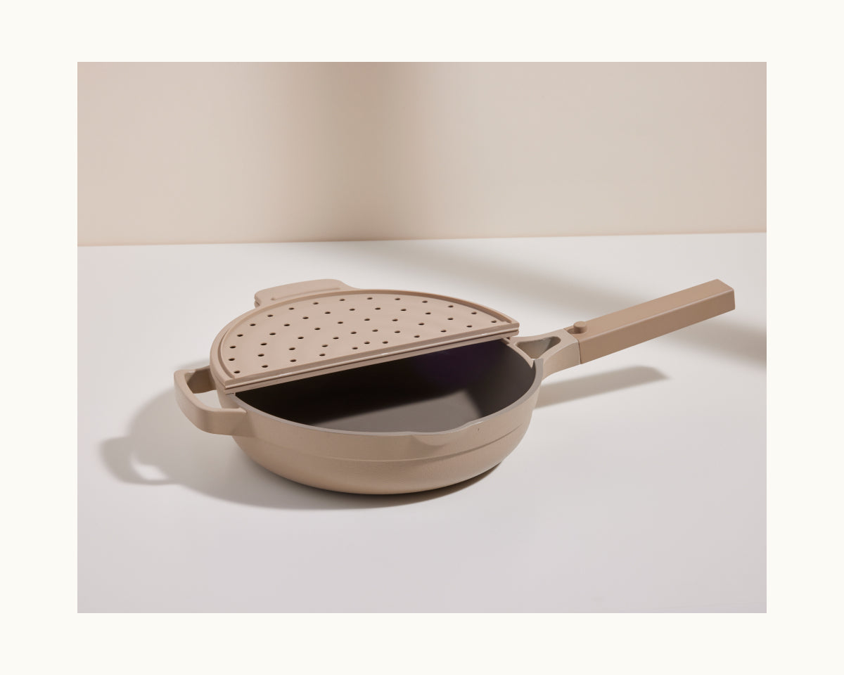 The Fearless Fry, a beige non-stick frying pan with a matching perforated lid for splatter prevention inspired by the Always Pan, rests on a white surface against a neutral background.