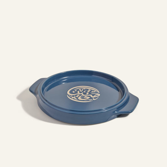 The Flipping Platter is a round blue ceramic server with side handles and a cream abstract lid design, ideal for serving dishes like tahdig or maqluba. Shown on a light background.