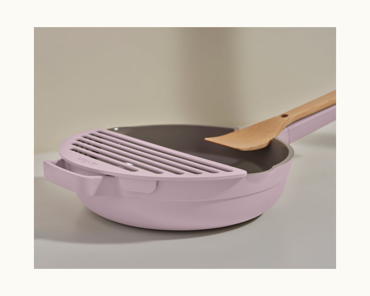 The Fry Deck, a matching light purple strainer attachment for the Always Pan from Our Place, is displayed with a wooden spatula on a neutral surface.