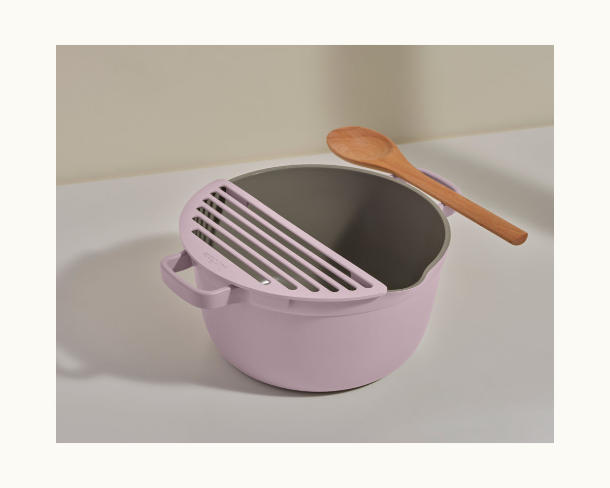 A light purple Fry Deck from Our Place, featuring a slotted lid attachment and a wooden spoon resting on top, is placed on a white surface.