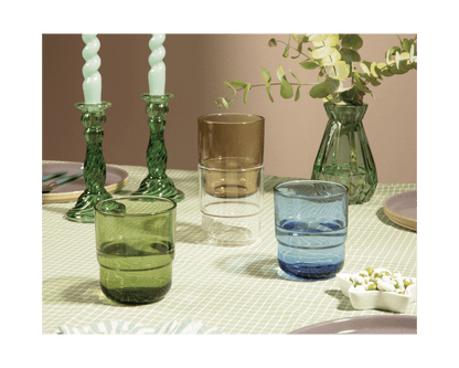 Three Short Night + Day Glasses in green, brown, and blue sit on a checkered table beside green candlesticks, candles, a vase with foliage, and a small dish.