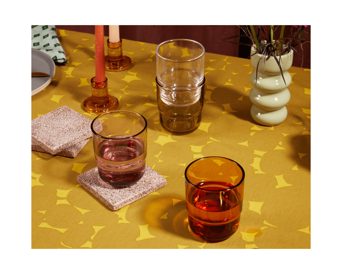 A yellow patterned tablecloth displays stacked and filled Short Night + Day Glasses in vibrant colors, two candle holders, stone coasters, a plant in a vase, and partially visible plates.