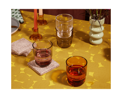 A yellow patterned tablecloth displays stacked and filled Short Night + Day Glasses in vibrant colors, two candle holders, stone coasters, a plant in a vase, and partially visible plates.