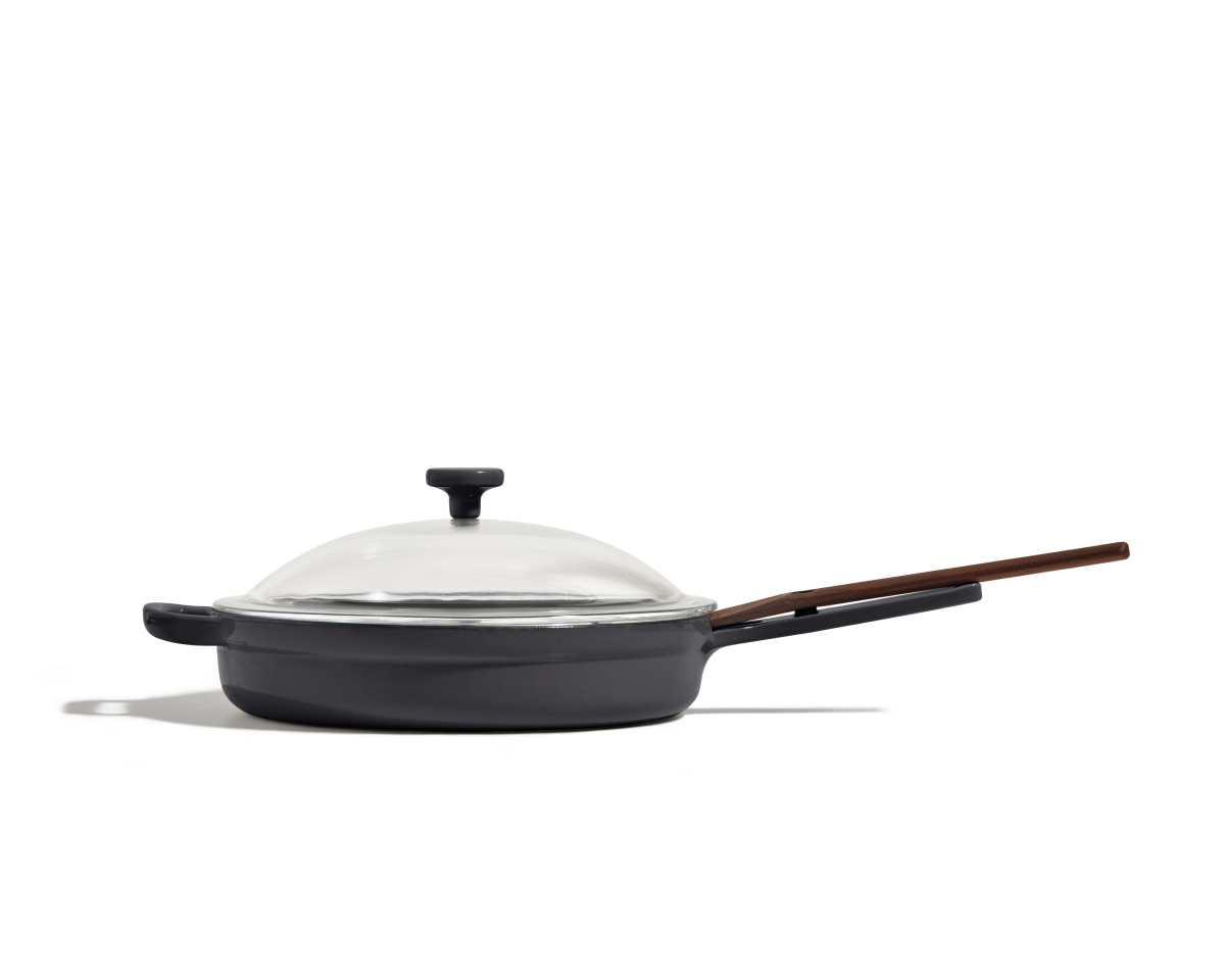 The Cast Iron Always Pan, featuring an enameled black finish, wooden handle, and clear glass lid, is shown from the side against a plain white background.