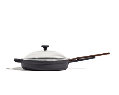 The Cast Iron Always Pan, featuring an enameled black finish, wooden handle, and clear glass lid, is shown from the side against a plain white background.
