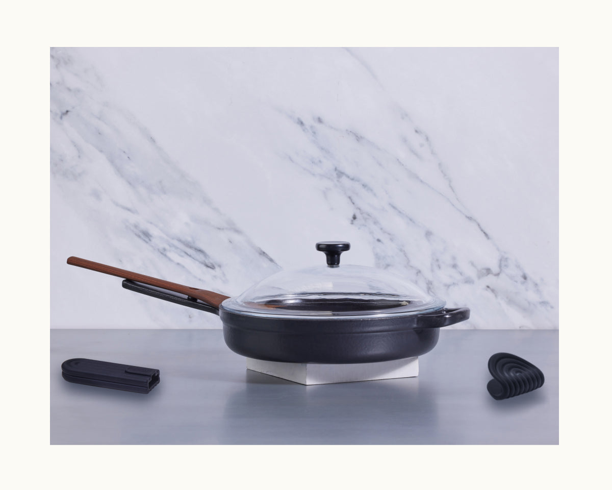 The Cast Iron Always Pan, featuring a black enameled finish, nonstick coating, glass lid, and wooden handle, sits on a metal surface with a black spatula and utensil rest nearby; marble backsplash in the background.