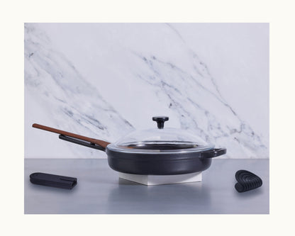 The Cast Iron Always Pan, featuring a black enameled finish, nonstick coating, glass lid, and wooden handle, sits on a metal surface with a black spatula and utensil rest nearby; marble backsplash in the background.