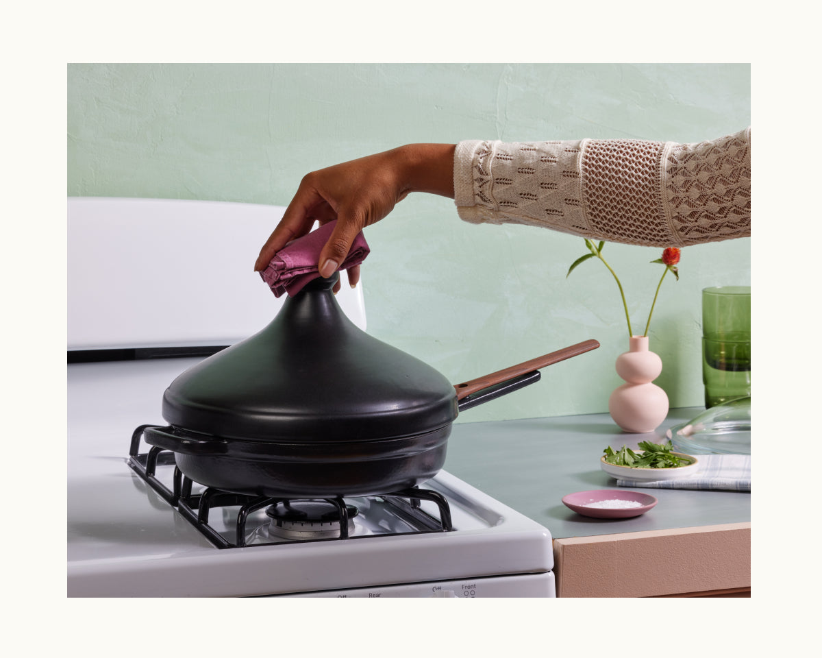 A person lifts the lid of a Cast Iron Always Pan on a gas stove using a cloth; nearby on the counter are a vase, bowl, and chopped herbs.