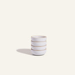 Four white Tiny Bowls, noted for their scratch-resistant ceramic finish, are stacked on a plain light background with a soft left shadow. These durable bowls are ideal as stylish and space-saving stackable tableware.
