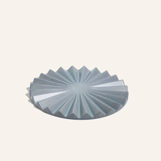 The Pleat Trivet is a round, blue-grey ceramic accessory featuring a geometric sunburst pleated pattern—ideal for protecting surfaces and adding decorative style to your kitchen.