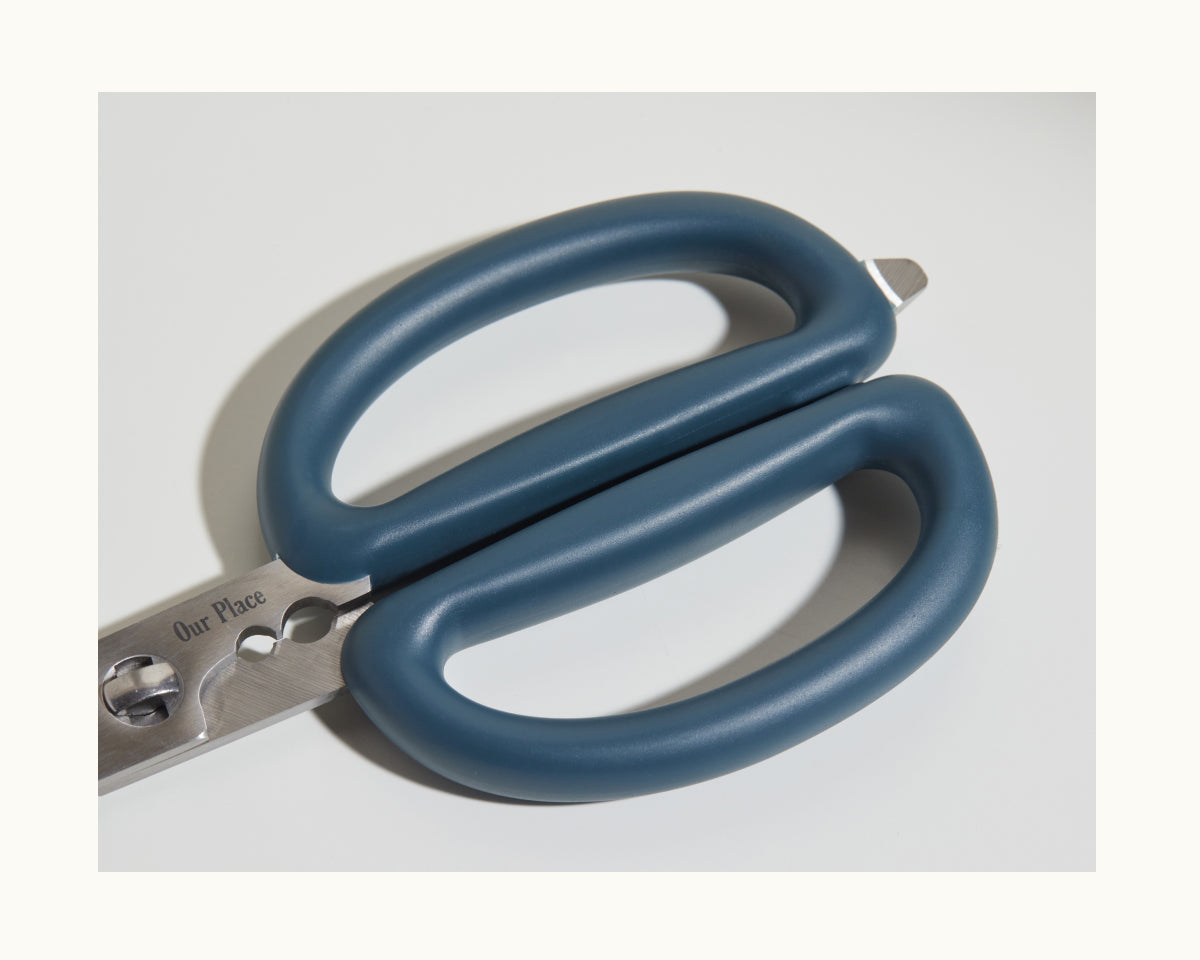 Close-up of the Shear Genius kitchen shears from Our Place, featuring blue handles, stainless steel blades, and a multifunctional design with built-in herb strippers.