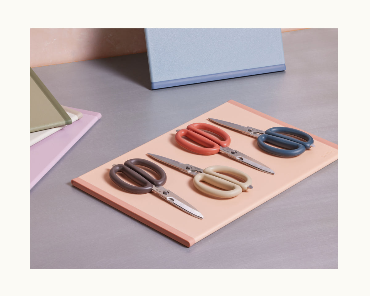 Four Shear Genius scissors with multicolored handles and a multifunctional design are displayed on a peach folder, surrounded by closed notebooks on a desk.