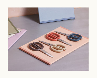 Four Shear Genius scissors with multicolored handles and a multifunctional design are displayed on a peach folder, surrounded by closed notebooks on a desk.