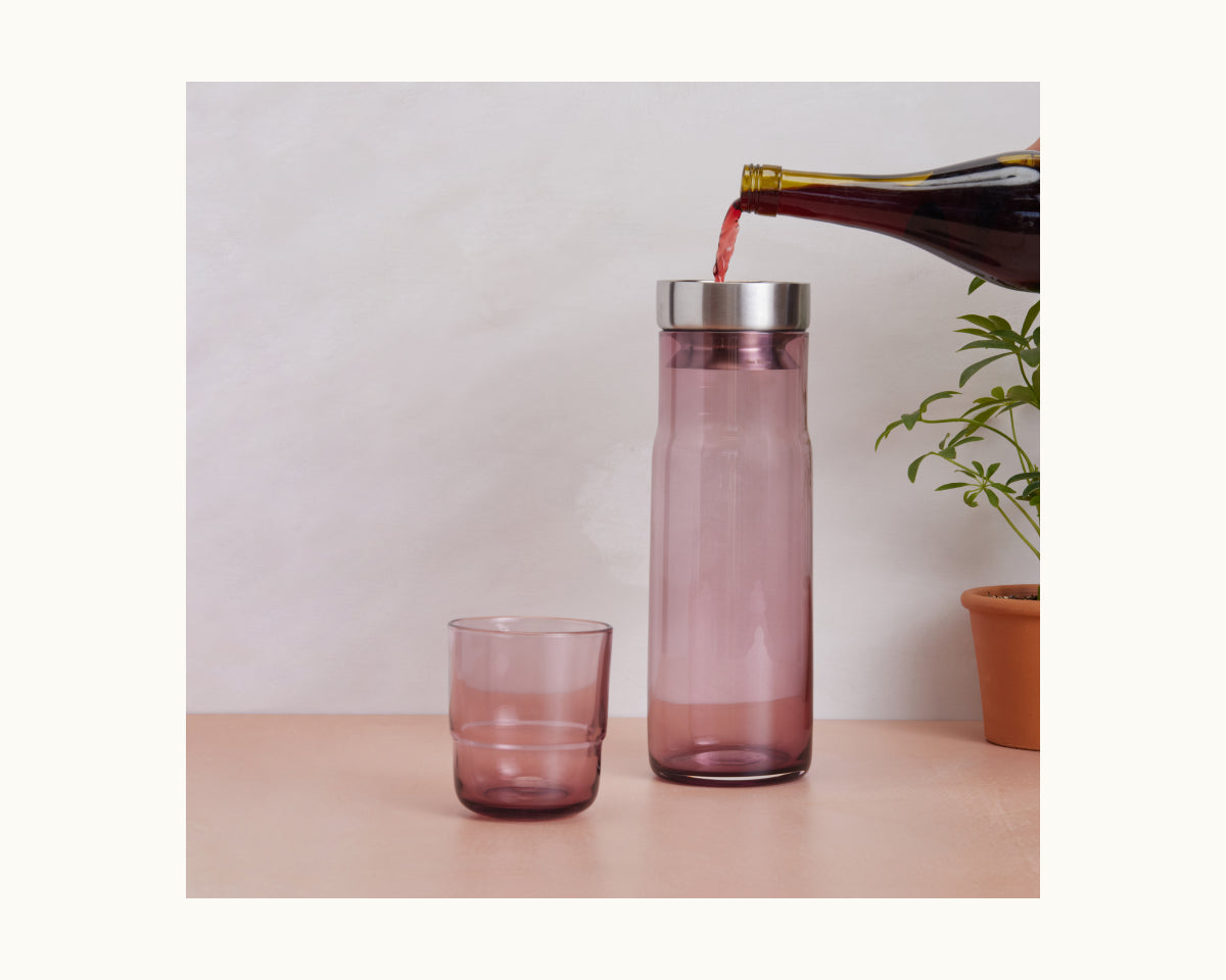 A dark liquid is poured from a bottle through a wine aerator into the Night + Day Carafe, which stands tall and purple beside a matching tumbler and a potted plant on a light surface.