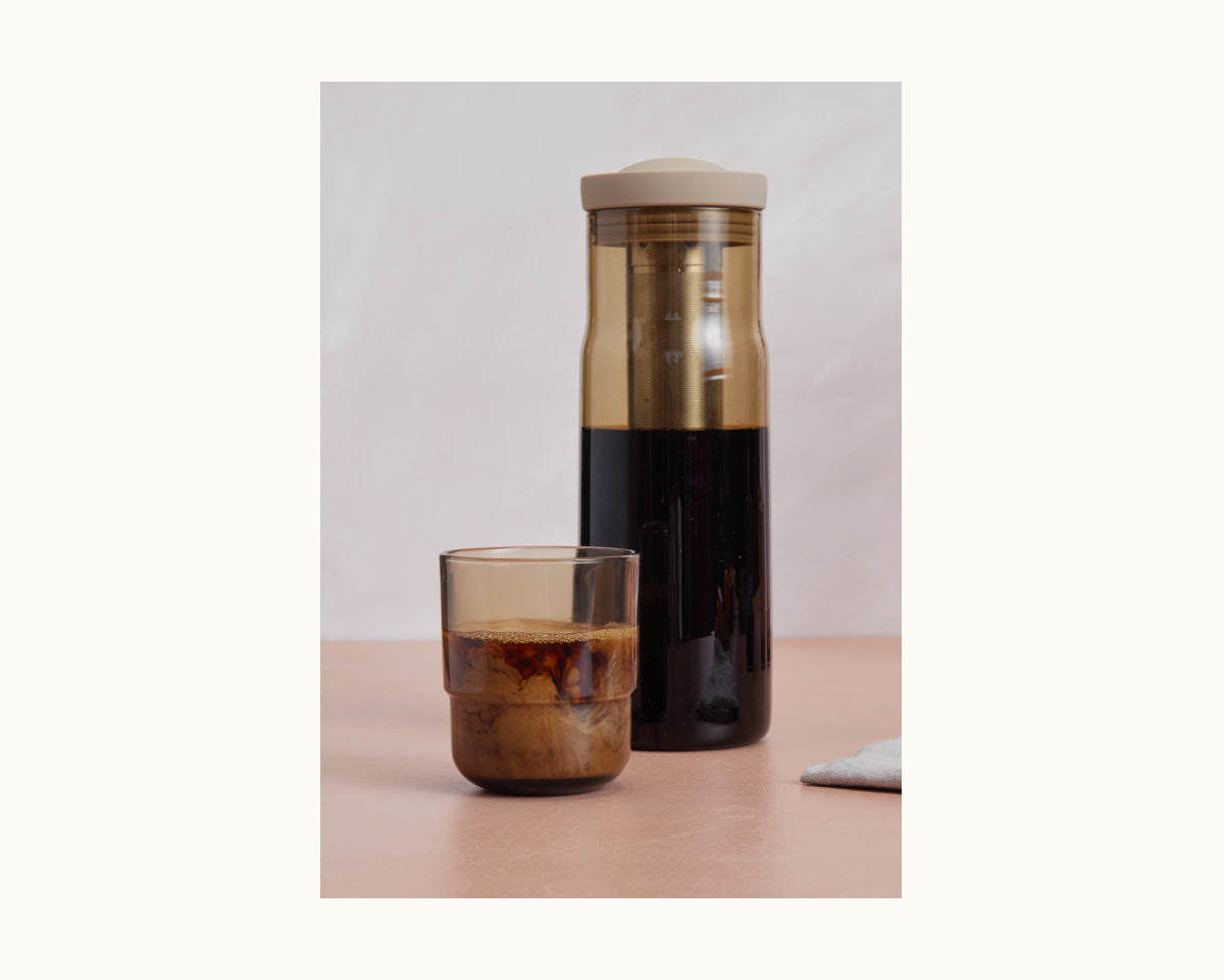A glass of iced coffee sits in front of the Night + Day Carafe cold brew infuser on a light surface with a white background.