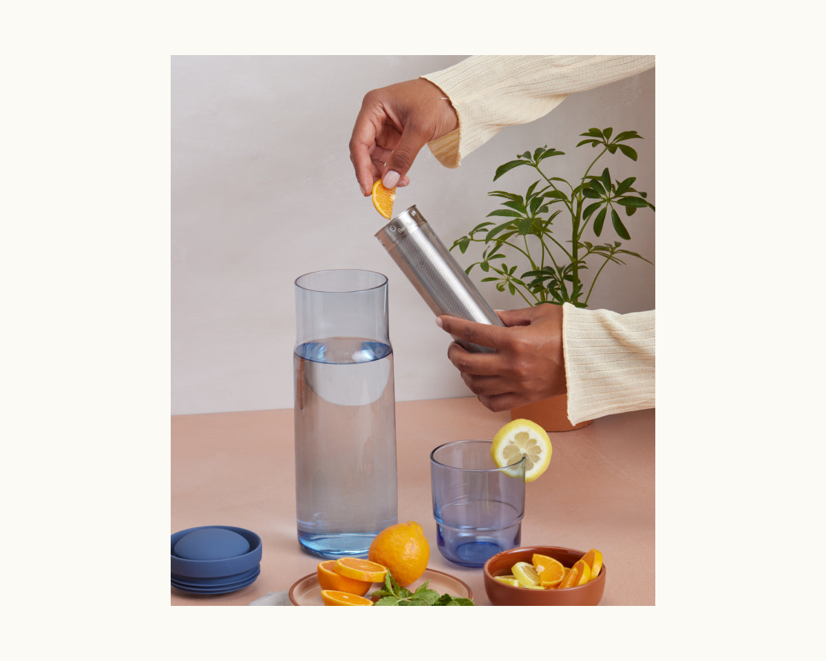 A person adds a lemon wedge to the Night + Day Carafe on a table with a glass, sliced lemons and oranges, blue lids, and a green plant in the background.