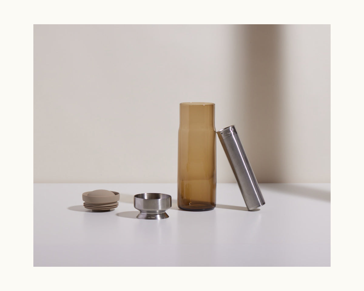The Night + Day Carafe set includes a brown glass bottle, metal cylinder, round metal cap, metal cup, and two beige disks displayed upright on a white surface against a neutral background.