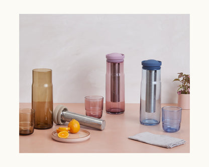 The Night + Day Carafe, two glass tumblers, three glass bottles with colored lids and filters, a plate of citrus, a gray cloth, and a small potted plant are displayed on a beige surface.