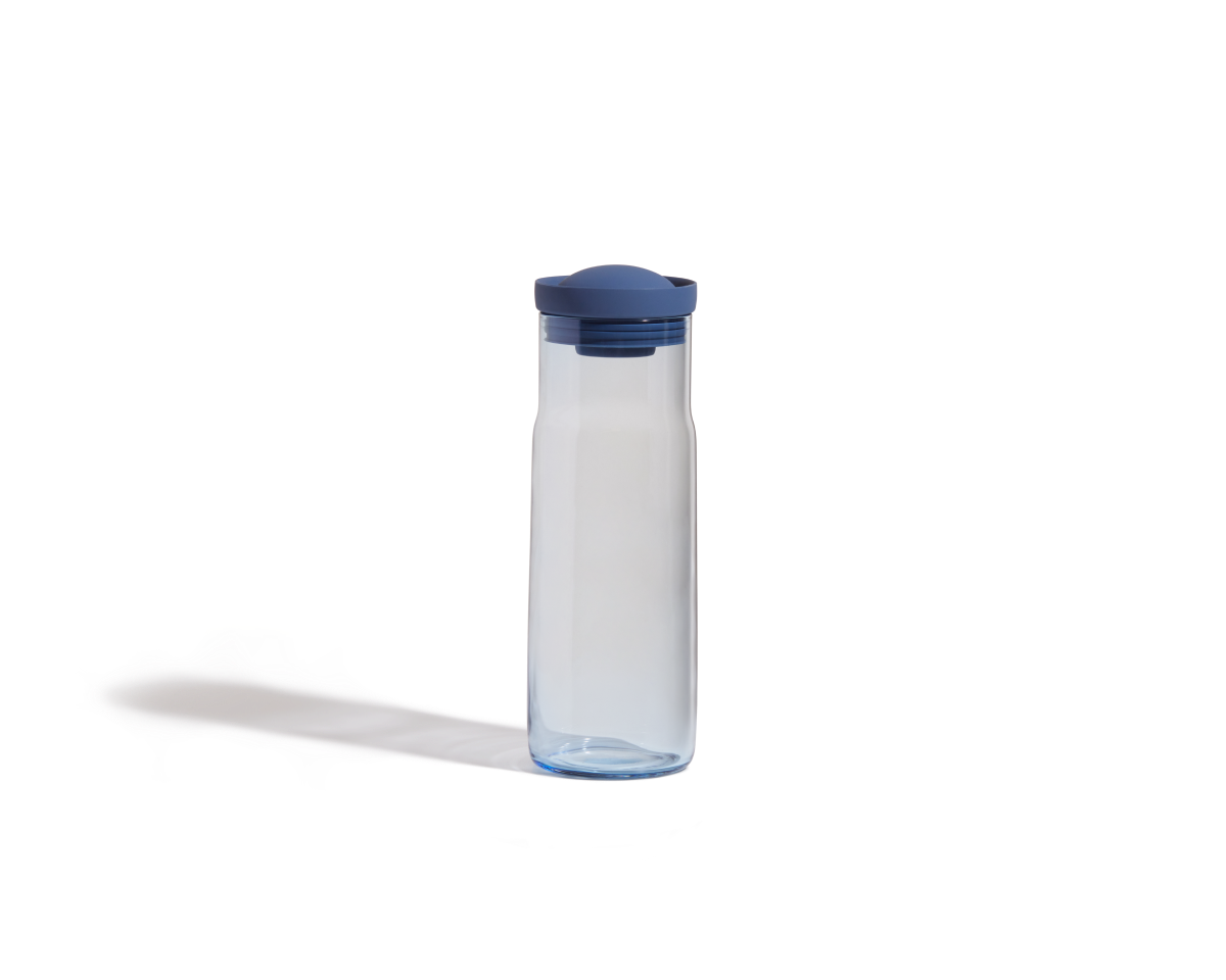 The Night + Day Carafe, a clear plastic bottle with a blue screw-on lid, stands upright on a white background and features a sleek, modern design.