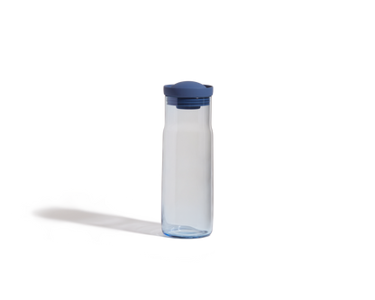 The Night + Day Carafe, a clear plastic bottle with a blue screw-on lid, stands upright on a white background and features a sleek, modern design.