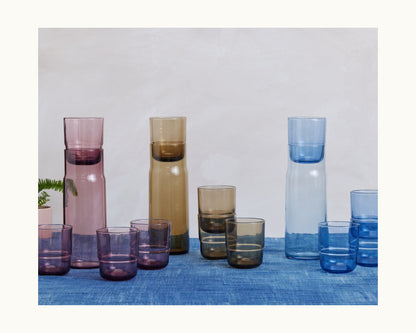 Three Night + Day Carafes with matching tumblers in pink, brown, and blue are arranged on a blue surface against a light background—ideal for use as a stylish wine aerator or cold brew infuser.