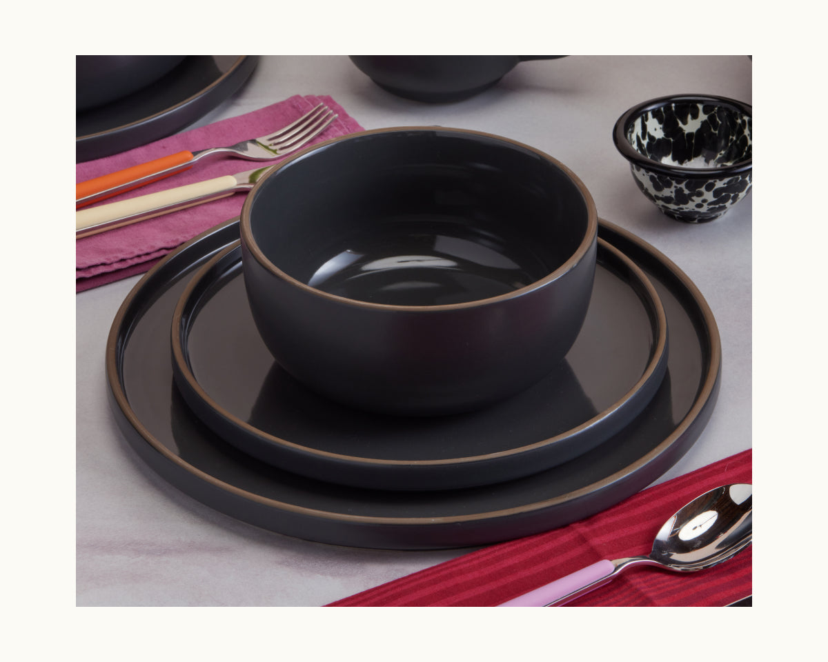 A Soup Bowl from a stylish ceramic set sits on a black plate and charger, accompanied by utensils, a small patterned bowl, and a spoon on a red napkin to complete the elegant tableware arrangement.