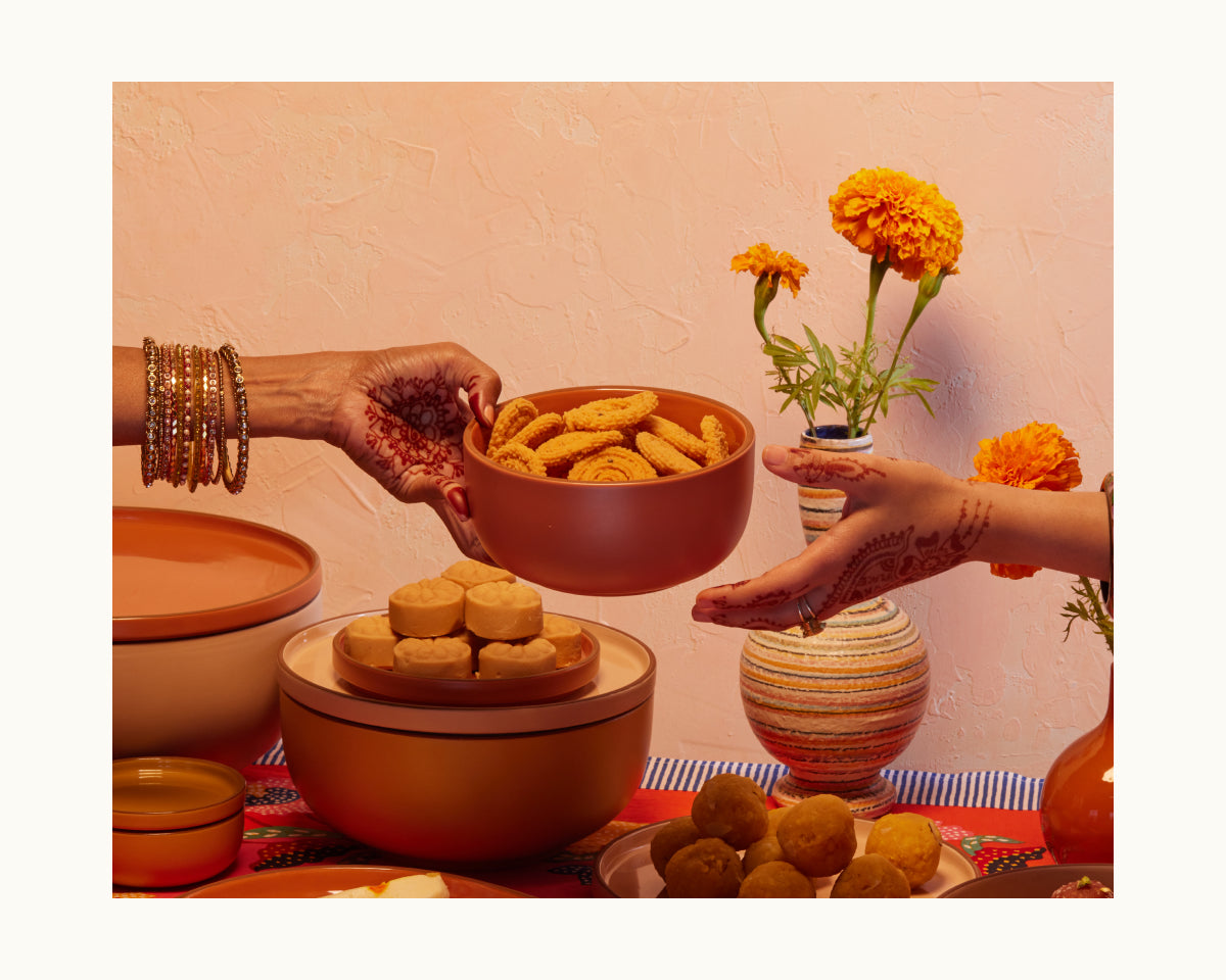 Two hands with henna and bangles exchange a bowl of Indian snacks over a table decorated with sweets, marigold flowers, and elegant Soup Bowls—perfect tableware for festive gatherings.