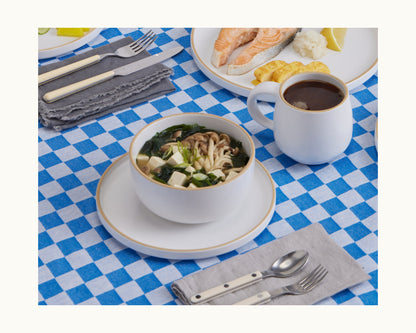 A bowl of noodle soup served in Soup Bowls, a mug of coffee, and a plate of assorted food are set on a blue and white checkered tablecloth with tableware and cutlery placed on gray napkins.