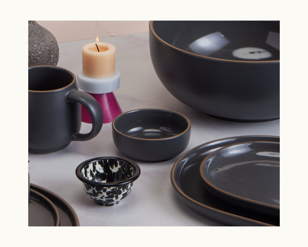 A set of stackable black ceramic Tiny Bowls and a matching mug arranged on a surface, with a candle and decorative cup in the background.